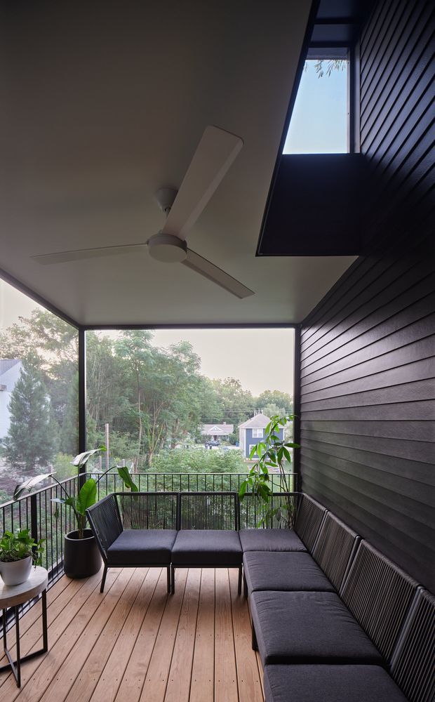 A contemporary covered outdoor balcony or terrace with a timber-plank deck, dark horizontal wall cladding, black metal-framed glazing and balustrades, and a ceiling fan. A dark upholstered L-shaped outdoor lounge and potted plants create a sheltered seating area overlooking leafy suburban surroundings.