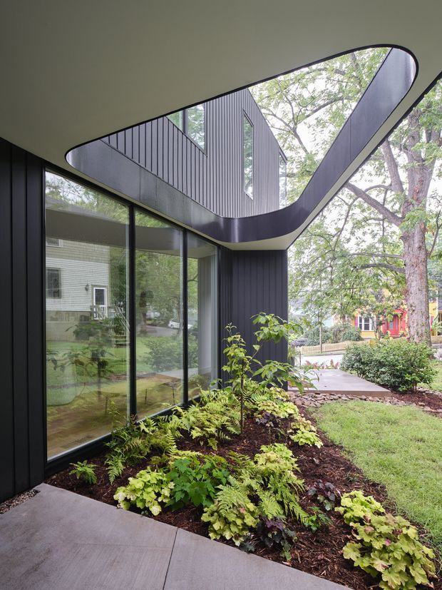 A contemporary residential extension viewed from an outdoor walkway, featuring dark vertically ribbed cladding, expansive floor-to-ceiling glazing, and a dramatic curved projecting roof or canopy forming an open sculptural edge. A planted garden bed with ferns and shrubs borders the concrete path, while mature trees and neighbouring homes are visible beyond.