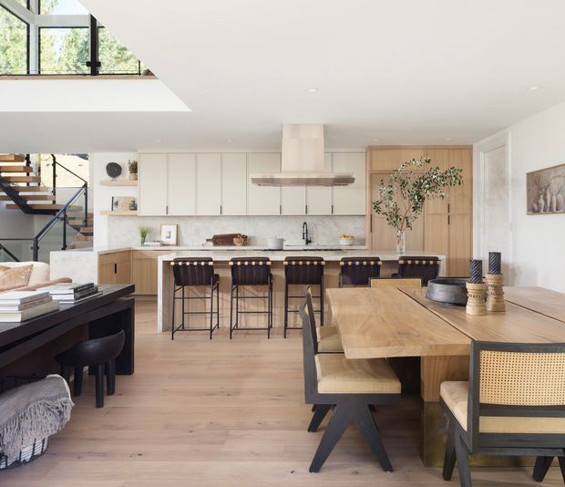 Bright open-plan interior combining a contemporary kitchen, dining area and living zone. The kitchen features white flat-panel cabinetry, a light stone benchtop, a large island with timber-and-black bar stools, and a sculptural rangehood. A substantial timber dining table with upholstered and cane-backed black-framed chairs occupies the foreground. Pale timber flooring, white walls, full-height timber joinery, recessed lighting and black metal stair detailing create a warm, refined modern look. A partial upper-level void and glazing are visible above the staircase.
