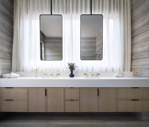 Contemporary bathroom vanity with a long double basin, pale timber cabinetry, white countertop, twin rounded-rectangle mirrors, and wall-to-wall sheer curtains. Grey wood-look wall and floor finishes create a calm, monochromatic scheme with dark metal mirror frames and tapware.