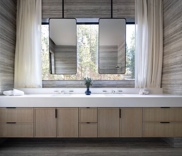 A refined contemporary bathroom vanity with a long double-sink counter, floating light-wood cabinetry, two rounded-rectangle mirrors, and floor-to-ceiling windows framed by sheer curtains. Grey wood-look wall and floor finishes create a cohesive, softly textured interior, with trees visible beyond the glazing.