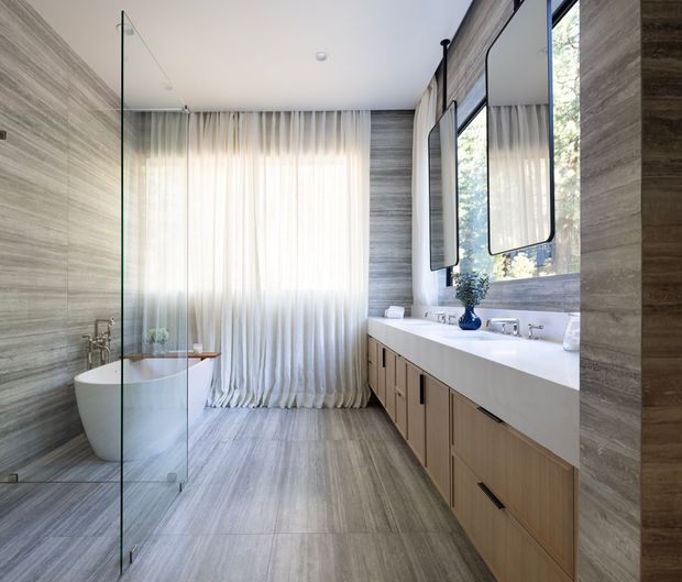 Bright, spacious contemporary bathroom with a freestanding white bathtub behind a clear glass shower screen, a long double vanity with timber-look cabinetry and a white countertop, large frameless mirrors, and grey wood-look tiled walls and floor. Full-height sheer curtains soften the natural light from the window.