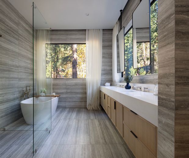 Contemporary bathroom with a freestanding white bathtub behind a frameless glass shower screen, a long double vanity with pale timber cabinetry and a white countertop, and large-format grey-brown stone-look wall and floor tiles. Full-height mirrors and a large window bring in natural light and views of trees outside.