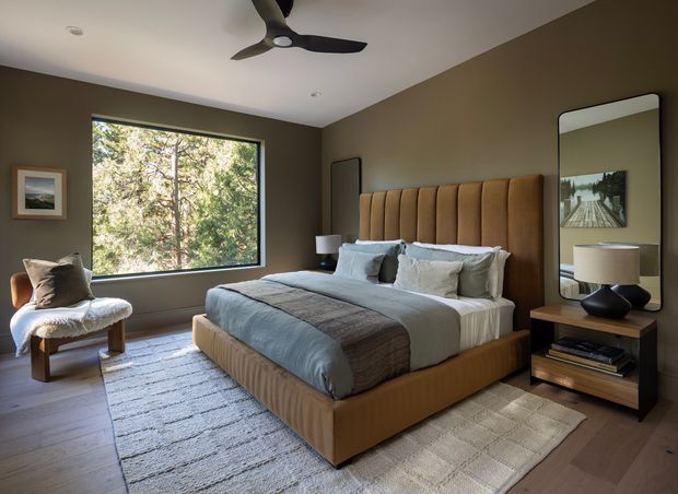 A refined contemporary bedroom with a large upholstered bed featuring a channel-tufted tan headboard and matching bed base, layered white and muted blue-grey bedding, and a textured area rug over timber flooring. Dark olive-brown walls, a large picture window overlooking trees, a sculptural ceiling fan, bedside lighting, a framed artwork, and a tall rounded rectangular mirror complete the warm, moody interior.