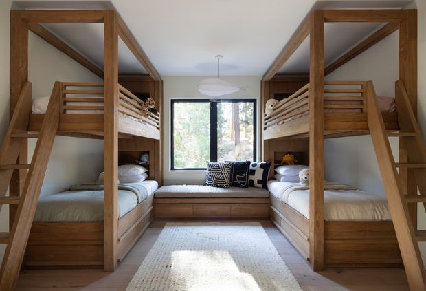 A compact shared bedroom featuring two pairs of built-in timber bunk beds facing each other, with integrated lower beds and upper bunks accessed by ladders. A central timber window seat sits beneath a large black-framed window overlooking trees, with neutral bedding, a patterned cushion, soft pendant lighting, and a pale rug. The room has a warm, practical, cabin-like character and abundant natural light.