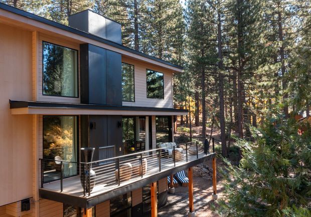 Exterior view of a contemporary multi-level house set among dense pine forest. The building has a compact flat or low-pitched roof form, light timber-toned cladding, dark metal wall panels, large windows, and a raised deck with horizontal metal balustrading supported on timber posts.
