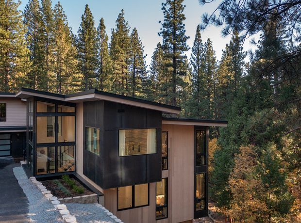 A modern multi-level forest dwelling set among tall pine trees. The building features dark vertical cladding, extensive floor-to-ceiling glazing, low-slope projecting rooflines, and a partially elevated form that follows the wooded terrain. A gravel-edged path and driveway lead toward the entrance.
