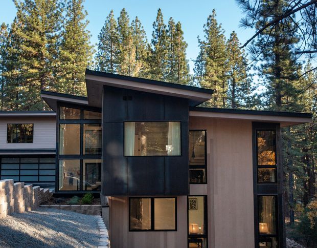 Exterior view of a modern multi-level house set among tall pine trees. The building features strongly projecting low-pitched roof planes, dark vertical cladding, pale rendered walls, extensive floor-to-ceiling glazing, and a stepped composition following a sloped, landscaped site.