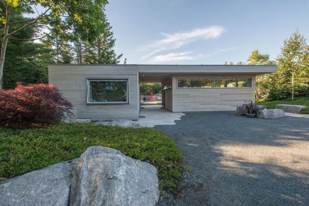 Single-storey modern house exterior with a low-pitched flat roof, horizontal grey wood cladding, large rectangular windows, and a covered recessed entry or carport-like opening. The building sits in a landscaped setting with mature trees, shrubs, boulders, and a dark gravel driveway.