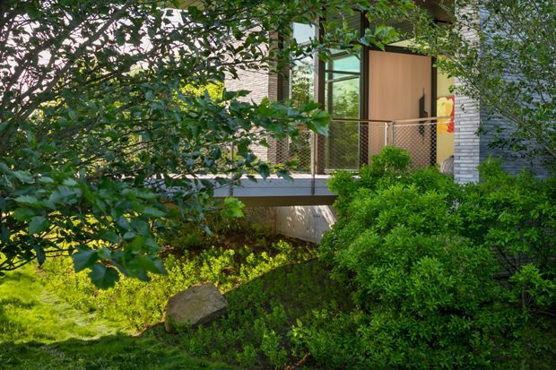 Exterior view of a contemporary residential entry or elevated walkway partially screened by mature greenery. The building features extensive glazing, a warm timber entry door, grey horizontal cladding, and a metal mesh balustrade over a raised platform. Dense shrubs, lawn, and landscaped planting surround the structure.