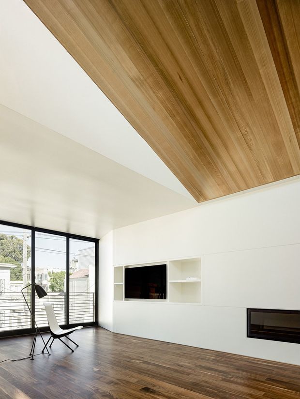 A bright, minimalist living room with full-height black-framed glazing, dark timber flooring, a sculptural lounge chair and floor lamp, white built-in media shelving, and a recessed linear fireplace. A prominent warm timber-lined ceiling feature adds contrast to the white walls and ceiling.