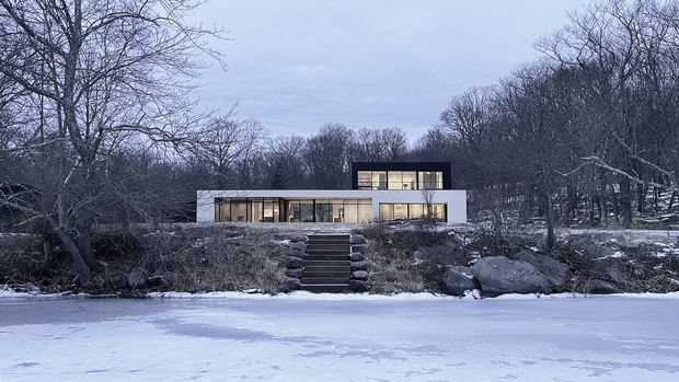 A modern, rectilinear two-storey house set among leafless trees on a snowy, frozen waterfront or lakeside site. The building has a low horizontal profile, pale rendered or masonry walls, extensive dark-framed glazing, and a projecting upper-level box. A central stairway leads up from the frozen foreground through a rocky, wooded landscape to the entrance.