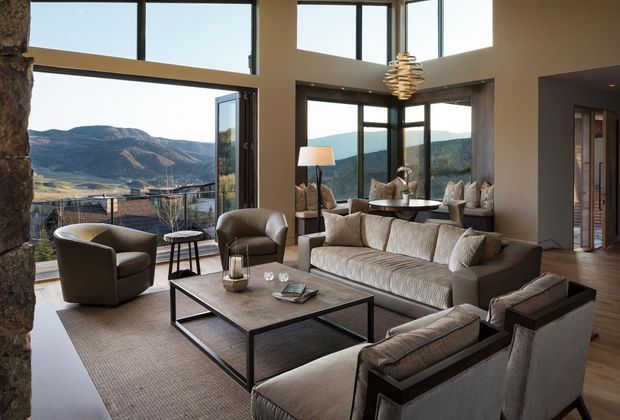 A spacious contemporary living room with floor-to-ceiling corner glazing overlooking a mountainous landscape. The room features a large neutral sectional sofa, two upholstered armchairs, a pair of additional lounge chairs in the adjoining seating area, a substantial rectangular coffee table, a textured area rug, timber flooring, and a sculptural pendant light. Natural stone is visible at the left edge, and an open doorway leads to another interior space.