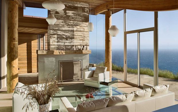 A coastal living room with expansive floor-to-ceiling glazing overlooking the ocean. The space features a large white sectional sofa, glass coffee table, exposed timber posts and ceiling, a stone fireplace with a weathered timber feature surround, sculptural pendant lights, and warm natural materials. The room is open, airy, and strongly connected to the landscape.