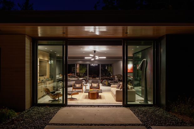 Night-time exterior view through a fully glazed opening into a warmly lit modern living room. The building has dark timber cladding, a timber-lined soffit, broad black-framed glass doors, and a simple paved entry path surrounded by dark gravel. Inside, a lounge seating arrangement, contemporary lighting, and a partially visible kitchen or bar area are visible.