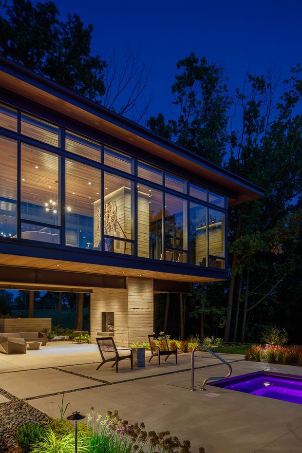 Twilight exterior view of a contemporary elevated house set among mature trees. The home features extensive floor-to-ceiling glazing, dark slender window frames, warm timber soffits and interior ceilings, and a cantilevered upper level supported by a stone-clad central volume. A paved outdoor terrace with lounge furniture, landscaped planting, and a small illuminated pool or spa occupies the foreground.
