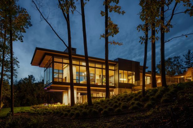 A modern multi-level house illuminated at dusk, set among tall trees on a sloping wooded site. The building features extensive floor-to-ceiling glazing, projecting flat roof planes, warm timber-toned soffits, dark cladding, and strongly lit interior spaces.