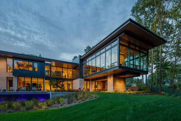 Exterior architectural photograph of a contemporary multi-level home set within a landscaped, wooded site at dusk. The design features strongly projecting flat roof planes, extensive floor-to-ceiling glazing, dark metal framing, warm timber soffits and wall cladding, and a cantilevered upper-level volume. Warm interior lighting creates a welcoming contrast with the cool blue evening sky and green lawn.