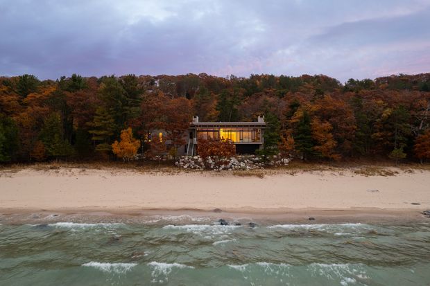 Aerial exterior view of a compact modern house set directly behind a sandy beach, surrounded by dense autumn-coloured woodland. The dwelling has a low, flat roof, extensive warm-lit glazing, and a rocky landscaped edge between the building and trees. Waves occupy the foreground under an overcast purple-blue sky.