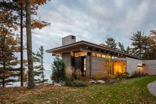 A low-slung contemporary house set among mature trees on a landscaped, sloping site. The exterior combines broad horizontal roof planes, exposed timber soffits, concrete walls and columns, dark-framed glazing, and warm timber-clad walls. Autumn foliage, ornamental grasses, stone landscaping, and a curving paved walkway surround the home. A glimpse of water or distant landscape is visible beyond the trees.