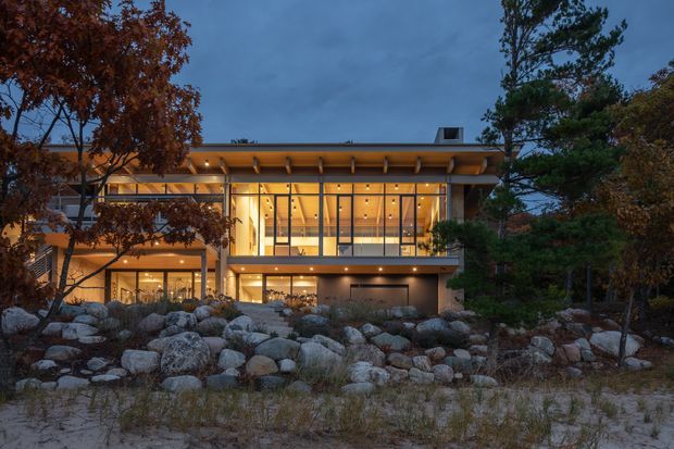 Twilight exterior view of a contemporary multi-level house set into a landscaped rocky site. The building features extensive floor-to-ceiling glazing, warm interior lighting, timber-toned soffits and framing, projecting horizontal rooflines, and a broad terrace or balcony. Large natural boulders, ornamental grasses, and mature trees form the foreground landscape.