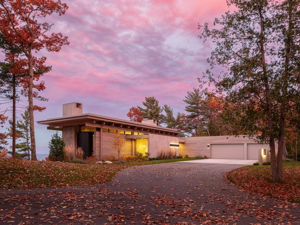 A low, single-storey modern residence photographed at dusk in an autumn woodland setting. The house features a broad flat roof with deep overhangs, warm timber-toned wall surfaces, clerestory-style glazing beneath the roofline, a recessed entry, and a separate integrated garage. A paved driveway curves through fallen leaves toward the home, while interior and exterior lighting create a warm glow against the pink-purple evening sky.