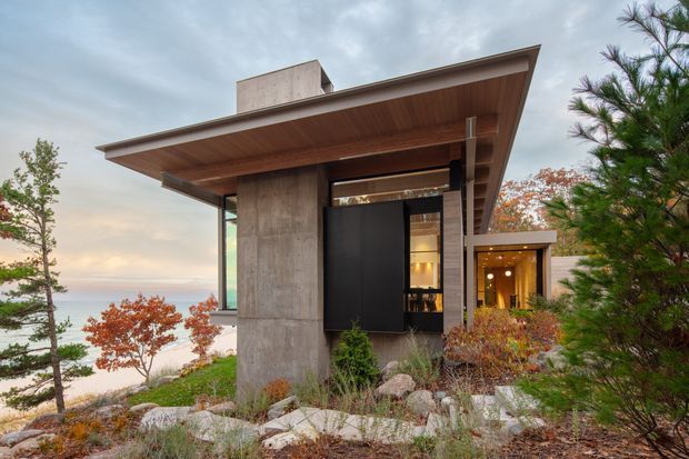 Exterior view of a contemporary, flat-roofed house set among landscaped rocky terrain and mature planting. The design features a broad cantilevered roof with a timber soffit, exposed concrete structural walls, dark framed glazing and a tall black entry door. Warm interior lighting is visible through the glazing, while the background suggests an elevated or waterside setting.