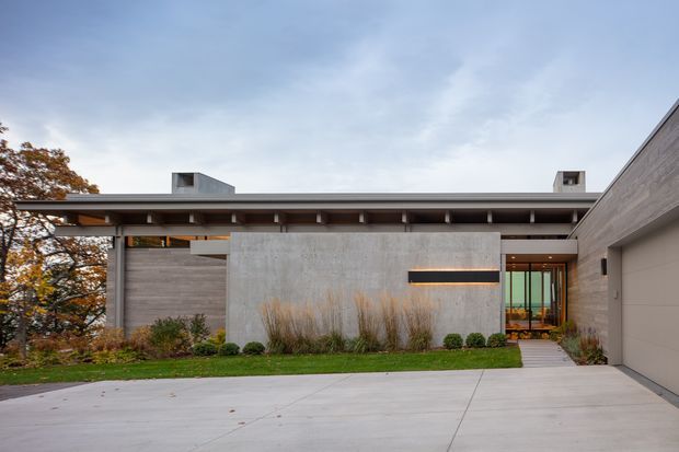 Single-storey contemporary residence viewed from the driveway, with a broad low-pitched roof and deep eaves, exposed dark structural beams, pale concrete or stone-clad walls, a central projecting feature wall with a horizontal recessed light, and glazed entry doors. A large pale driveway and integrated garage occupy the foreground and right side, with restrained landscaping around the facade.