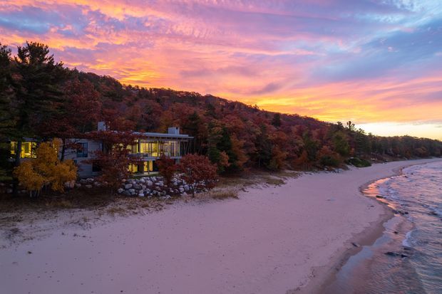 A modern multi-level residence nestled into a wooded hillside beside a sandy beach, photographed at sunset. The home has extensive illuminated glazing, low horizontal forms, and appears integrated with the rocky, forested coastal site. The beach and calm shoreline dominate the foreground, beneath a dramatic pink, orange, and purple sky.