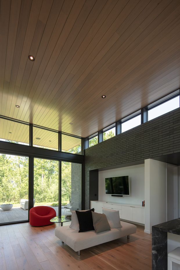 A contemporary double-height living room with a warm timber-lined ceiling, expansive black-framed glazing, dark brick feature walls, and timber flooring. The room contains a low white sofa, a vivid red sculptural lounge chair, a glass coffee table, and a wall-mounted television set within white built-in joinery. A kitchen island or benchtop is partially visible in the foreground, while a leafy outdoor area can be seen through the glazing.