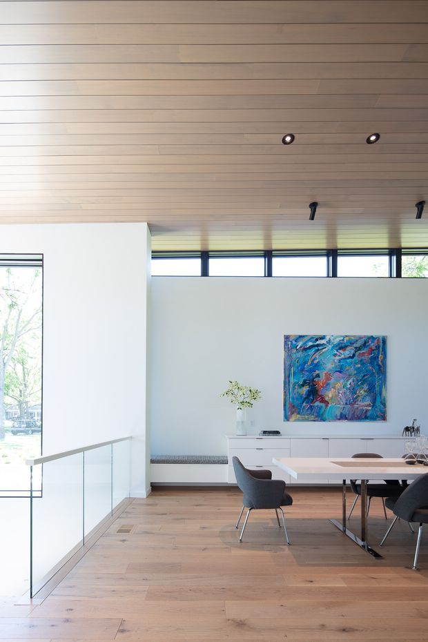 Bright contemporary dining area with a large timber-lined ceiling, pale timber flooring, white walls, and expansive clerestory and full-height glazing. A long light-toned dining table with dark upholstered chairs sits beside a streamlined white built-in sideboard, colourful abstract artwork, and a glass balustrade overlooking a lower level. Recessed and directional ceiling lights provide understated illumination.