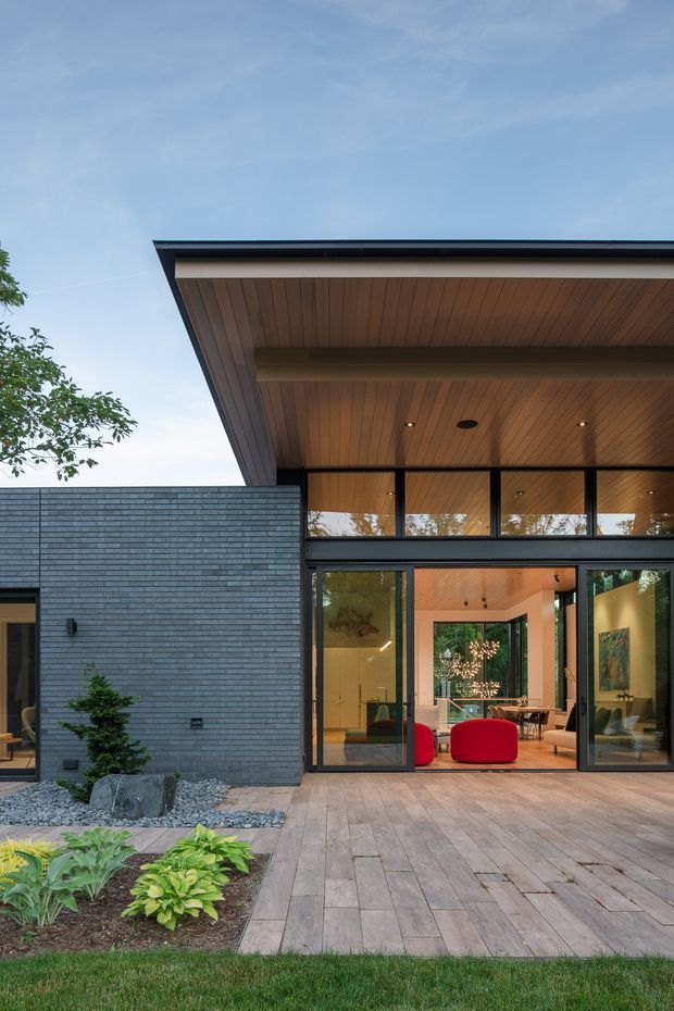 Exterior view of a contemporary single-storey home opening onto a timber deck and landscaped garden. The facade combines dark horizontal brickwork with expansive black-framed glazing, while a broad projecting roof has a warm timber-lined soffit with recessed lighting. The open doors reveal a refined living and dining area with colourful seating and timber finishes.