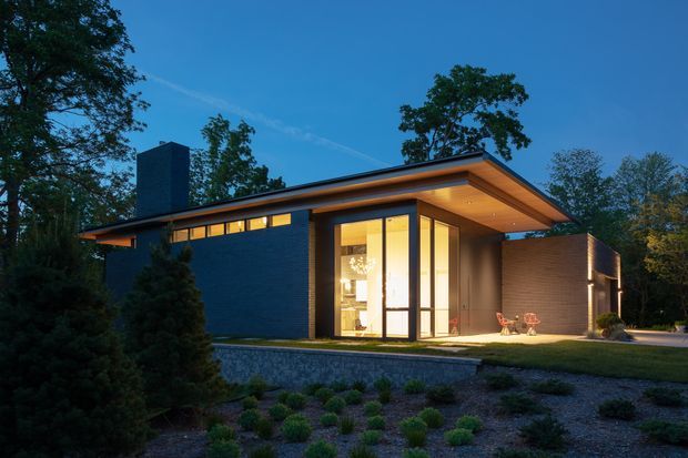 Twilight exterior view of a low-slung contemporary house set within a landscaped, wooded site. The home features a broad flat roof with deep overhangs, dark masonry walls, warm timber soffits, extensive floor-to-ceiling glazing, and a tall chimney-like vertical volume. Interior lighting creates a warm focal point through the glazed entry or living area.