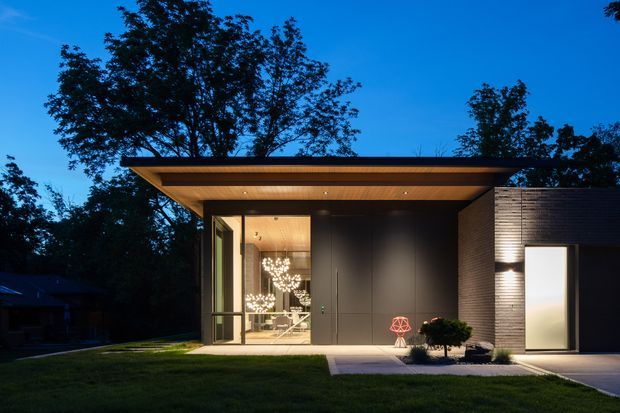 Twilight exterior view of a contemporary single-storey pavilion-like residence. The building has a broad flat projecting roof with a warm timber soffit, dark smooth wall panels, textured horizontal masonry cladding, and expansive glazed openings revealing a warmly lit interior. A small landscaped lawn, paved entry path, and carefully placed exterior lighting frame the facade.