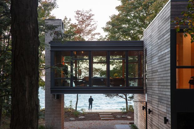Exterior view of a contemporary multi-level residence set among mature trees beside a body of water. A dark-framed glazed bridge-like upper volume connects or spans between masonry and shingle-clad sections, with a warm-lit interior visible through the windows. A gravel path and steps lead toward the waterfront.