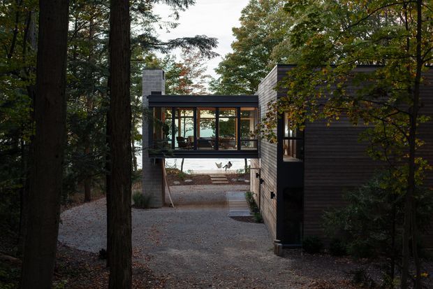 Exterior view of a modern, dark-clad house nestled among mature trees. The composition shows a compact glazed connection or bridge between building volumes, a flat-roofed glass enclosure, a pale masonry chimney, dark horizontal wall cladding, and a gravel approach through the wooded site.
