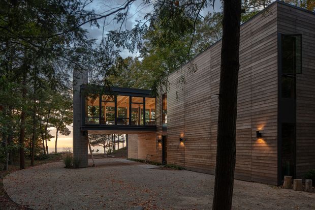 Exterior view of a contemporary timber-clad house set among mature trees. The building has a long horizontal wing with a glazed elevated living area, dark-framed windows, a projecting flat-roofed volume, and warm exterior wall lighting. A gravel forecourt and surrounding woodland frame the structure, with a glimpse of water or sky beyond the trees.