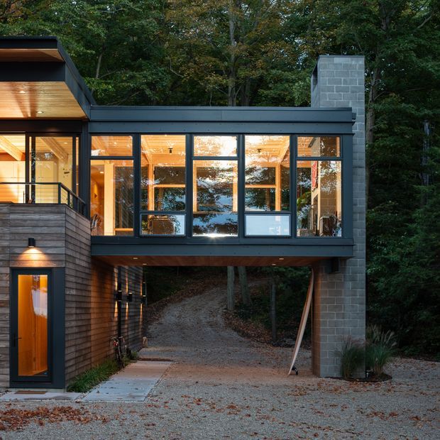 Exterior view of a contemporary, elevated house set within a wooded landscape. The building combines dark horizontal cladding, extensive black-framed glazing, warm timber soffits and interiors, and a tall exposed concrete-block chimney or side wall. A covered driveway or passage runs beneath the upper-level living space, with an entry door and balcony visible at left.