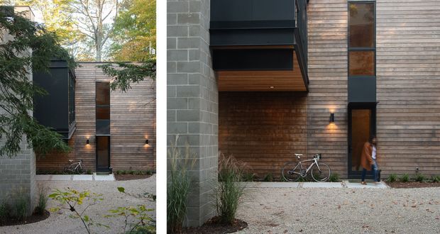 A split-view exterior photograph of a contemporary multi-level residence. The facade combines horizontal timber weatherboard cladding, dark metal-framed projecting window or balcony elements, exposed grey concrete block walls, and warm brickwork around a recessed entry. Gravel paving, restrained landscaping, bicycles, and wall-mounted lighting frame the entrance courtyard.