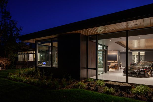 Twilight exterior view of a low-slung contemporary house with extensive floor-to-ceiling glazing, dark cladding, and a broad flat roof overhang. Warm timber soffits and interior lighting contrast with the dark facade, while a polished garage or showroom-like space with vehicles is visible through the glass. The building is set within landscaped garden beds.