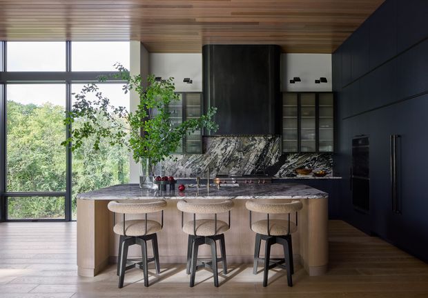 A sophisticated contemporary kitchen with a large marble island and three stools, dark navy cabinetry, a dramatic veined stone splashback, integrated appliances, and a warm timber-lined ceiling. Floor-to-ceiling glazing brings in abundant natural light and views of greenery.