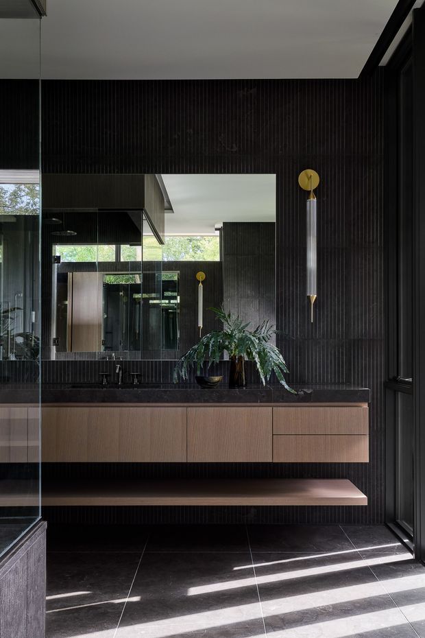A sophisticated contemporary bathroom with a long floating timber vanity, dark stone benchtop, integrated twin basins and black wall-mounted tapware. The room is lined with dark vertically ribbed wall panels and matching dark floor tiles, with a large mirror, full-height glazing, a built-in bench, and sculptural gold wall lights. Natural light creates strong linear shadows across the floor, while a large leafy plant adds an organic contrast.