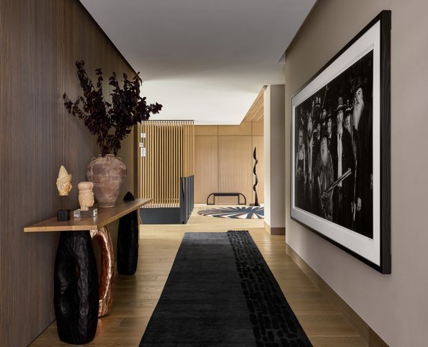 A refined residential hallway or entry gallery with warm timber flooring, a dark vertically textured timber feature wall, and a slim console displaying sculptural objects. A large black-and-white framed artwork dominates the right wall. The corridor leads toward a brighter open area with vertical timber screening, a bench, and additional contemporary artwork. The scheme is restrained, warm, and monochromatic with strong sculptural accents.