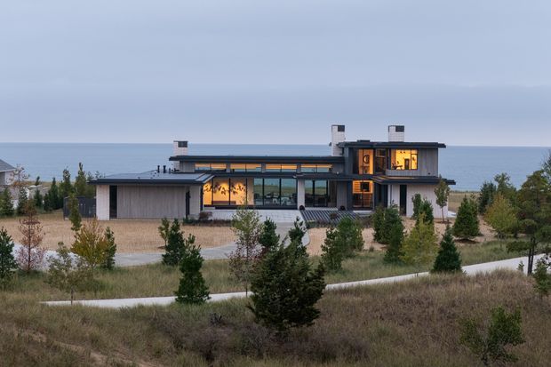 A modern two-storey coastal house viewed from landscaped grounds, with a low horizontal composition, flat rooflines, extensive glazing, dark metal framing, and warm interior lighting. The property sits beside a visible body of water, surrounded by natural grasses, young trees, and curved paved paths.