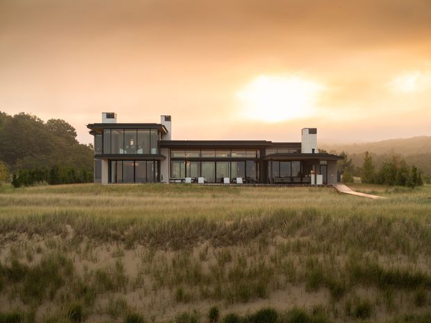 A wide exterior view of a contemporary multi-level home set within a grassy, lightly wooded rural or semi-rural landscape. The building has a long, low horizontal form with extensive floor-to-ceiling glazing, dark framed windows, flat or very low-pitched roof planes, projecting upper-level volumes, and prominent chimney elements. A narrow walkway leads from the house into the surrounding grassland. Warm sunset light creates a muted, atmospheric scene.