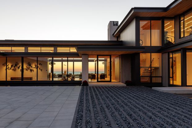 Contemporary multi-wing residence viewed from a paved courtyard at dusk. The home features strong horizontal and rectilinear volumes, extensive floor-to-ceiling glazing, dark metal-framed windows and doors, warm timber soffits and cladding, pale masonry walls, and a textured linear stone driveway or paving treatment. Warm interior lighting glows through the glazing, revealing living and circulation spaces.