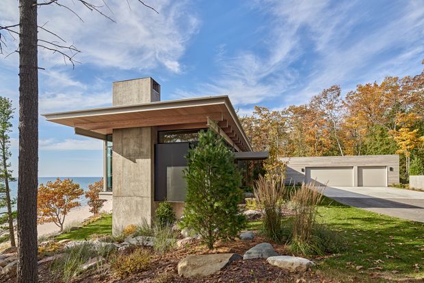 Exterior view of a low-slung modern home set in a landscaped, wooded site beside a body of water. The house features a broad shallow roof overhang, exposed warm timber soffit, tall concrete wall elements, dark-framed glazing, and a detached or connected flat-roofed garage. Autumn trees, lawn, rocks, and a driveway surround the building.
