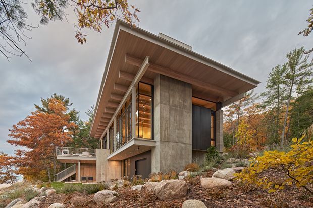 Exterior architectural photograph of a contemporary multi-level house set into a wooded, rocky landscape. The building features a broad low-pitched roof with a deep timber-lined soffit, exposed projecting rafters, extensive floor-to-ceiling glazing, concrete feature walls, dark vertical cladding or panels, and a projecting balcony. Autumn trees and large natural boulders surround the structure.