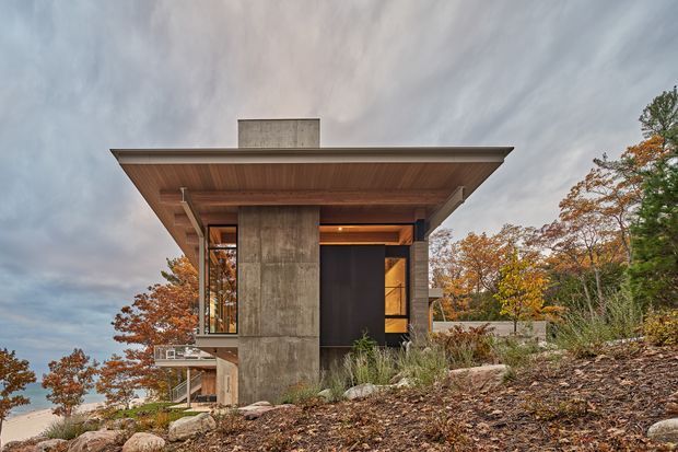 Exterior view of a contemporary, flat-roofed house set into a wooded sloping site. The compact structure features a broad cantilevered roof with a timber-lined soffit, a tall exposed concrete central wall or chimney volume, dark-framed glazing, and warm interior lighting. Autumn trees, native-style grasses, rocks, and the elevated terrain surround the building.