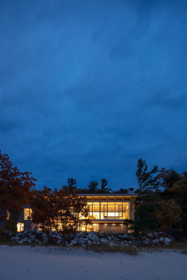 Twilight exterior view of a modern, flat-roofed multi-level house set behind a rocky landscaped edge. The building has extensive warm-lit floor-to-ceiling glazing, slim dark frames, projecting horizontal roof and balcony lines, and is surrounded by mature trees and large boulders. A sandy or pale ground plane occupies the foreground.